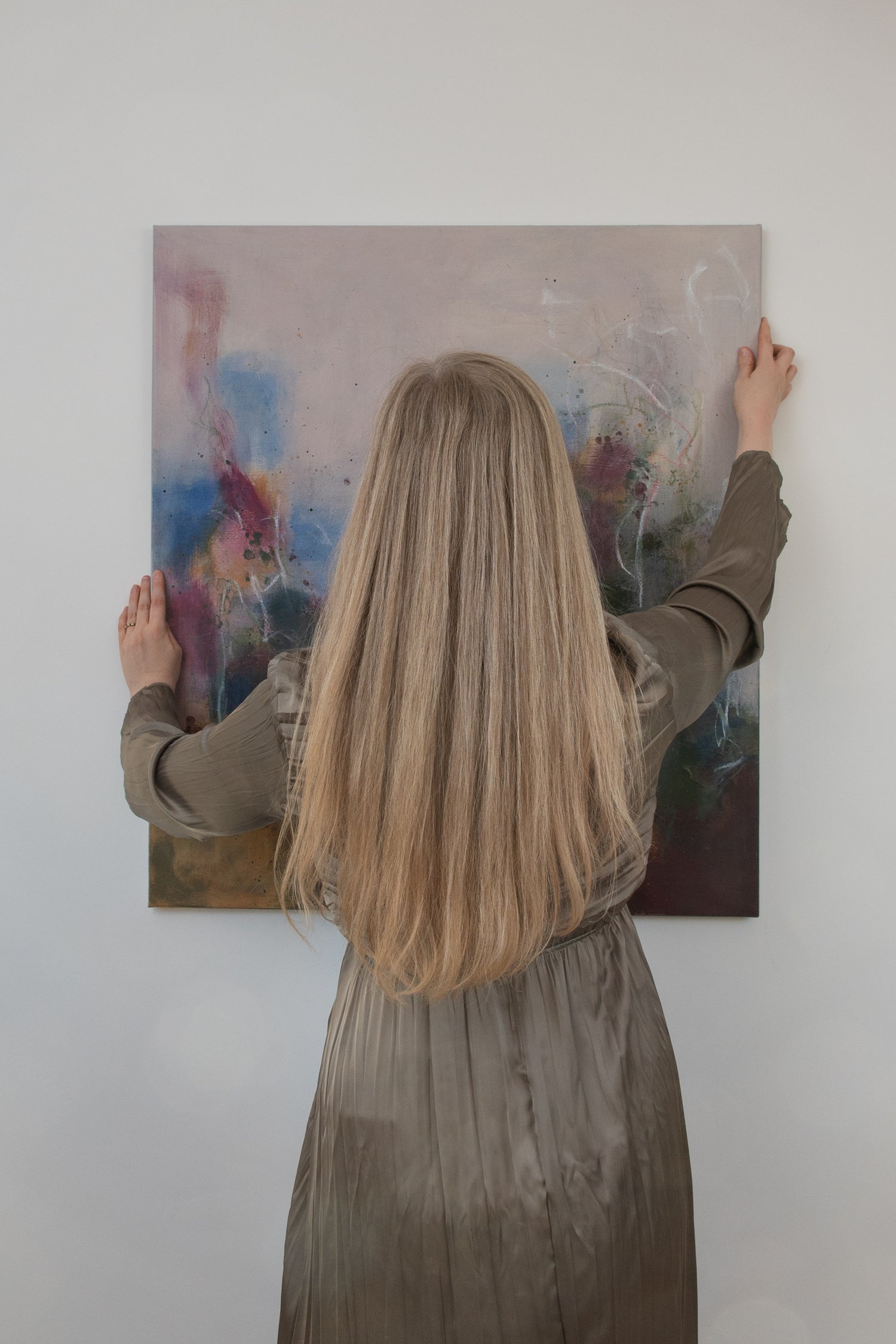 Bethany, a woman with long blond hair in a green dress, is seen from behind holding a painting up to the wall, to be hung.