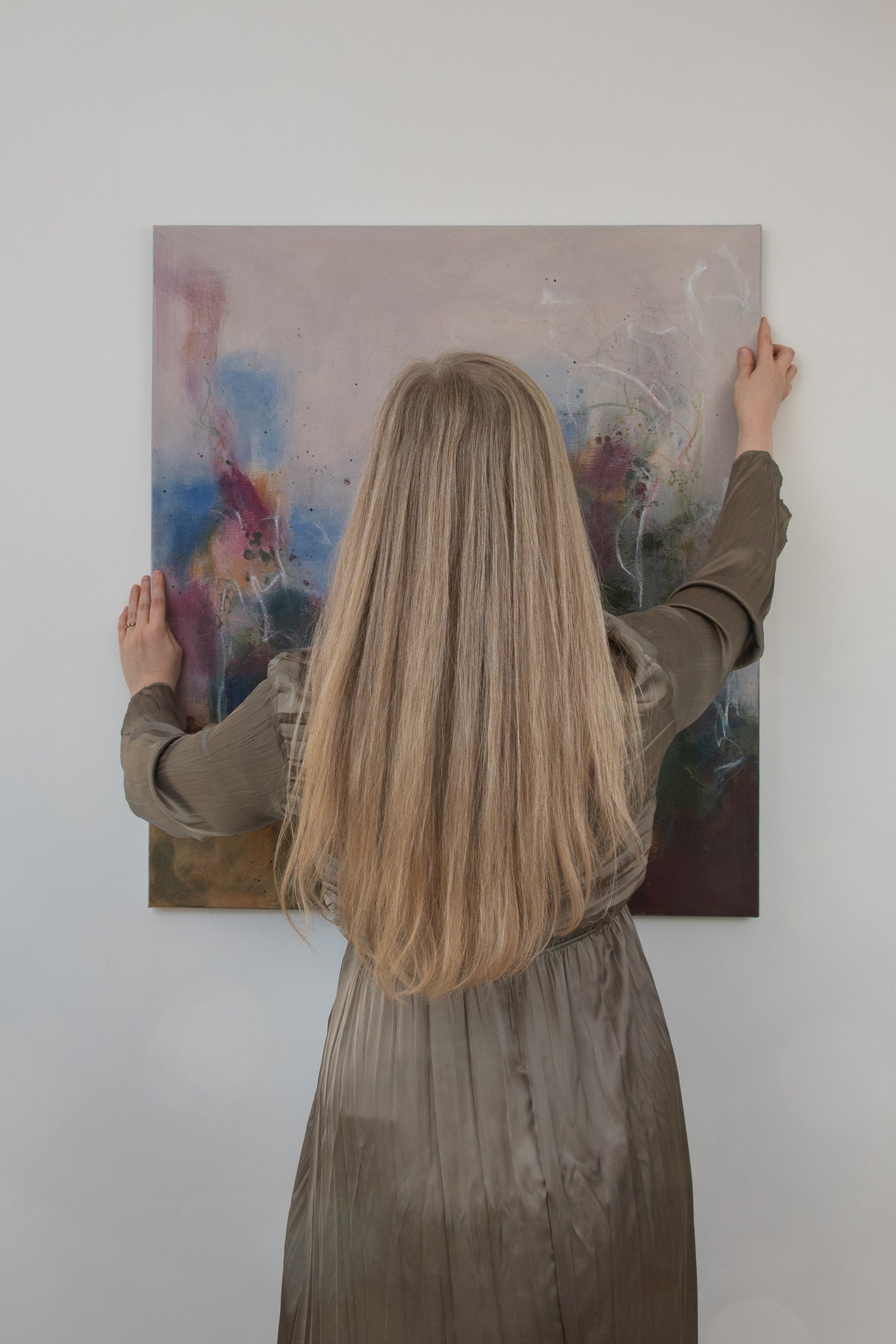 Bethany, a woman with long blond hair in a green dress, is seen from behind holding a painting up to the wall, to be hung. 